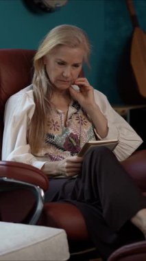 Çekici orta yaşlı bir kadının portresi canlı bir sandalyede oturmuş günlüklerini okuyor, güzel dekore edilmiş bir mekanda rahat bir anın tadını çıkarırken gülümsüyor..