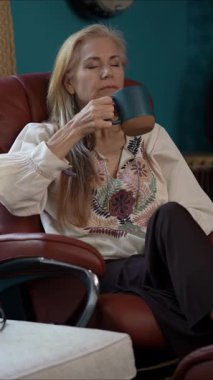 Orta yaşlı, sarı saçlı bir kadının rahat bir sandalyede oturup deftere yazdığı dikey bir video. Sıcak ve davetkar bir atmosferle çevrili, odaklanmış ve rahat görünüyor..