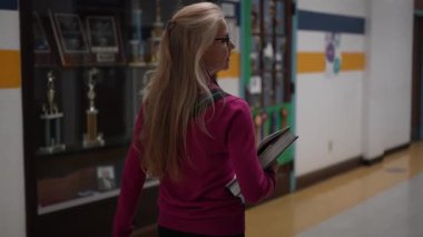 Pembe kazak giymiş orta yaşlı bir kadın okul koridorunda yürür, elinde kitaplarla kupa vitrinine bakar..