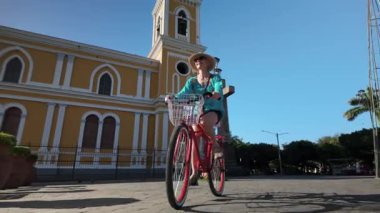 Mutlu, gülümseyen bir kadın, açık mavi gökyüzünün altındaki güzel bir katedralin yanında Granada, Nikaragua sokaklarında bisiklet sürmekten zevk alır..