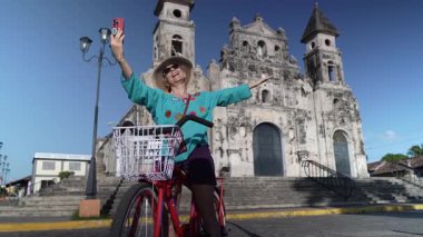 Olgun bir kadın akıllı telefonuyla Granada, Nikaragua 'daki tarihi Iglesia Guadalupe' nin önünde bisiklet sürerken selfie çeker ve seyahat deneyiminin tadını çıkarır..