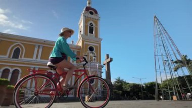 Mutlu bir kadın bisikletiyle Nikaragua, Granada 'nın canlı caddelerinde yürüyor, arkasında heybetli bir katedral yükseliyor, neşeli bir seyahat deneyimi sergiliyor..