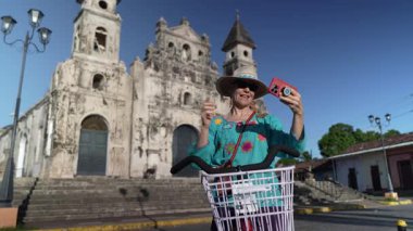 Gülümseyen bir kadın, Granada, Nikaragua 'da tarihi Iglesia Guadalupe kilisesinin yanında telefonu ve bisikletiyle selfie çekiyor. Mutluluğunu açık hava deneyimini kucaklayarak ifade eder..