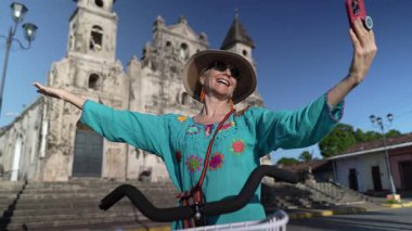Mutlu bir kadın, Granada, Nikaragua 'da bisiklete binmekten keyif alırken, tarihi Iglesia Guadalupe Kilisesi' nin önünde açık bir gökyüzünün altında telefonuyla selfie çekiyor..