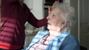 Bakıcı, yaşlı bir kadının saçlarını parlak ve rahat bir odada tarar. Kadın bir sandalyede dinleniyor, öğleden sonra şımartma anının tadını çıkarıyor..