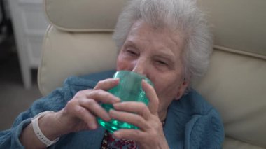 Yaşlı bir kadın bir bardak alıp bir yudum su içerken rahat bir yaşam alanında, huzur ve neşe saçar..