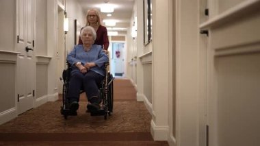 Bakıcı, yaşlı bir kadına tekerlekli sandalyede iyi aydınlatılmış bir huzurevinin koridorunda rehberlik eder, bağlarını ve günlük aktivitelerini vurgular..