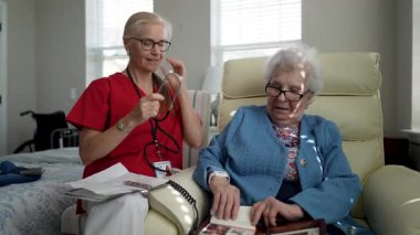 Bir hemşire, bir fotoğraf albümüne bakarken rahat bir şekilde oturan yaşlı bir kadının sağlığını dikkatle kontrol ederek bakım ve bağlantının önemini vurgular..