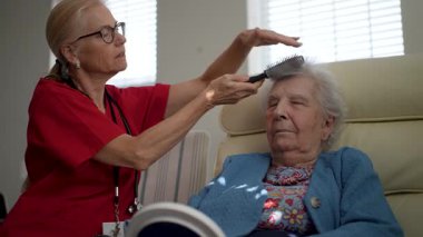 Bakıcı, yaşlı bir kadının saçlarını nazikçe şekillendirir. O da rahat bir şekilde sandalyede oturur. Güneş ışığı pencereden süzülerek sıcak bir atmosfer yaratır..