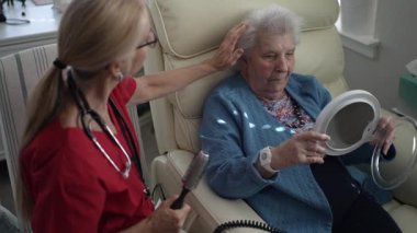 Bakıcı, yaşlı bir kadına el aynasında yansımasını incelerken saçı için nazikçe yardım eder. Sıcak, davetkar atmosfer onların etkileşimini güçlendirir..