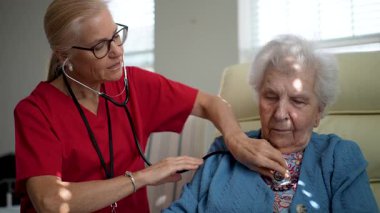 Bakıcı yaşlı bir kadının kalp atışlarını dikkatle dinler. Rahat bir ortamda, şefkat ve sağlık hizmetlerine olan bağlılığını gösterir..