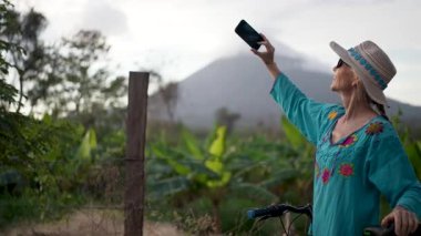 Renkli kıyafetli, gülümseyen olgun bir kadın, Nikaragua 'daki Ometepe Adası' nda Concepcion Volkanı 'nda bisiklet sürerken telefonuyla fotoğraf çekiyor. Neşeli macerasının tadını çıkarıyor..