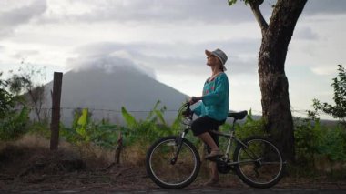 Gülümseyen olgun bir kadın Nikaragua 'daki Ometepe Adası' nda bisiklet sürüyor. Concepcion volkanı bulutlu bir gökyüzünün altında görkemli bir şekilde yükseliyor..