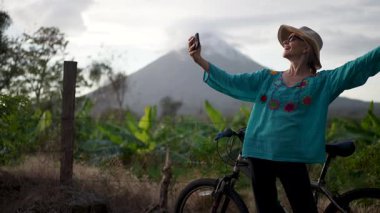 Renkli bluzlu mutlu bir son sınıf güzeli, Ometepe Adası 'nın arka planında yemyeşil bir yeşil ile çevrili Concepcion volkanı ile neşeli bir selfie çeker..