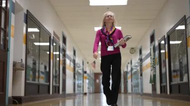 Orta yaşlı bir kadın, şık giyinmiş, okul koridorunda elinde kitaplarla geziyor, derslere hazırlanıyor..