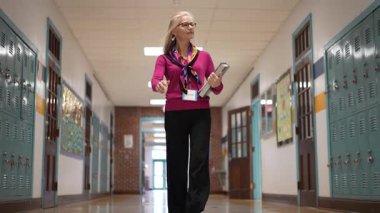 Berkeley Springs, WV, ABD - 125 2024: Gözlüklü ve renkli eşarplı orta yaşlı bir kadının portresi okul koridorunda yürüyor, kitap taşıyor ve arkadaşça ve ulaşılabilir bir ışık saçıyor