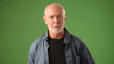 Olgun bir adam kollarını kavuşturup yeşil bir zemine karşı düşünceli bir ifade sergiliyor. Kıyafeti onun seçkin varlığına katkı sağlıyor..