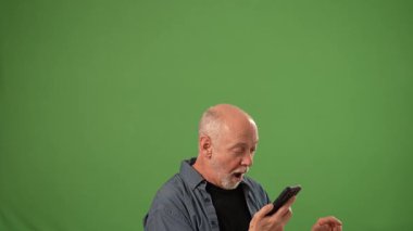 Neşeli bir ifadeye sahip olgun bir adam akıllı telefonunu yukarı doğru tutar, sanki yeşil bir arkaplan önünde heyecan verici haberleri paylaşır gibi..