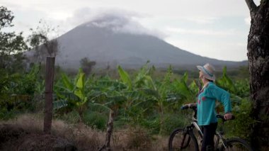 Geniş açı, güzel yaşlı bir kadın Omotepe Adası 'nda yemyeşil bir araziyle çevrili bisiklet sürüyor. Concepcion volkanı arka planda görkemli bir şekilde beliriyor.