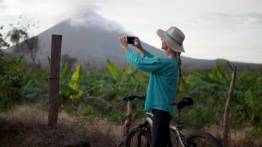 Mutlu bir son sınıf öğrencisi Nikaragua, Ometepe 'de bisiklet sürerken telefonuyla fotoğraf çekiyor. Arka planda görkemli Concepcion volkanı yükseliyor..