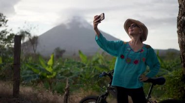 Renkli bluzuyla gülümseyen çekici yaşlı bir kadının Ometepe Adası 'nın arka planında yemyeşil çimenliklerle çevrili Concepcion volkanı ile mutlu bir selfie çekmesi..