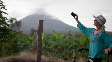 Renkli kıyafetli, gülümseyen olgun bir kadın, Nikaragua 'daki Ometepe Adası' nda Concepcion Volkanı 'nda bisiklet sürerken telefonuyla fotoğraf çekiyor. Neşeli macerasının tadını çıkarıyor..