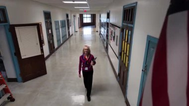 Berkeley Springs, WV, ABD - 125 2024: Olgun bir öğretmen okul koridorunda Amerikan bayrağıyla güvenle yürüyor. Eğitimsel bir törende güne hazırlanan bir klasörü tutarken dolaplar duvarlara dizilir.