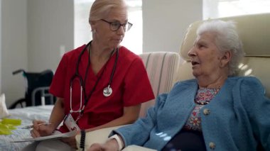 Kırmızı önlüklü bir bakıcı mavi kazaklı yaşlı bir kadınla iletişim kurar, sağlık hizmetlerinde nişan ve konforu teşvik eder..