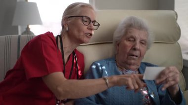 Bakıcı yaşlı bir kadınla iletişim kurar, hatıraları canlandırmak için bir fotoğrafa işaret eder. Bağlantılar, sağlık merkezlerindeki kişisel bakım merkezlerini vurguluyor.
