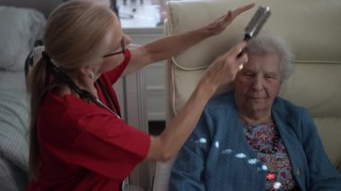 Bakıcı, yaşlı bir kadının saçını hafifçe tarar, güneşli bir öğleden sonra sıcak bir oturma odasında rahatlık ve bakım sağlar..