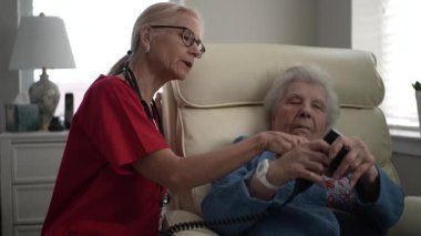 Kırmızı önlüklü bir sağlık çalışanı, özel bakım ve ilgiyi vurgulayarak, rahat bir yaşam alanında bir cihazla oturan yaşlı bir kadına yardım ediyor..