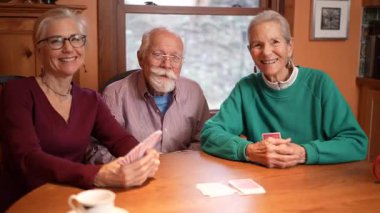 Üç aile üyesinin portresi, sıcak bir mutfak masasında kağıt oynarken keyifli bir anı paylaşıyor. Pencereden süzülen güneş ışığı gülümsemelerini ve kahkahalarını vurguluyor..