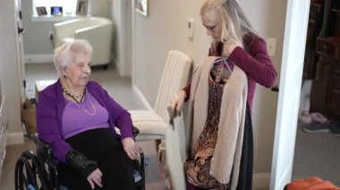 Bakıcı, rahat bir ev ortamında kendine kıyafet seçerken tekerlekli sandalyedeki yaşlı bir kadına yardım eder. Bu an özen ve desteği yansıtır..