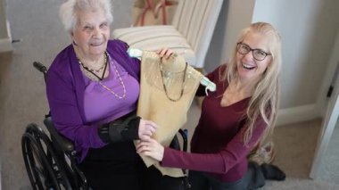 Tekerlekli sandalyedeki yaşlı kadın portresi, bir bakıcı ile ışıl ışıl bir kıyafet tutarken gülümsüyor. Parlak ve rahat bir ortamda keyifli bir anı paylaşıyorlar..