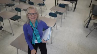 Orta yaşlı mutlu bir kadın, elinde dostane bir gülümsemeyle bir yığın kağıtla bir sınıfta duruyor. Masalar boş ve doğal ışık boşluğu dolduruyor..