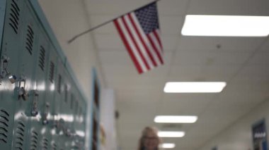 Orta yaşlı, gözlüklü ve mavi eşarplı bir kadın, elinde dolaplarla dolu bir okul koridorunda elinde kitaplarla odak noktasına doğru yürür ve gülümseyerek arkadaşça bir öğretim ortamı sunar..