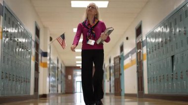 Berkeley Springs, WV, ABD - 125 2024: Yetişkin bir kadın öğretmen okul koridorunda güvenle yürüyor, kitap taşıyor. Salonda renkli dolaplar ve bir Amerikan bayrağı var..