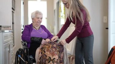 Bir bakıcı, rahat ve bakımlı bir odada otururken tekerlekli sandalyedeki yaşlı bir kadına kıyafetlerini ayarlamasında yardımcı olur..