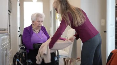 Tekerlekli sandalyedeki olgun bir kadın, iyi ışıklandırılmış iç mekandaki kıyafetlerini düzenleyen bir bakıcıdan yardım alır. Etkileşimleri samimiyet ve özen gösteriyor..