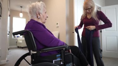 Tekerlekli sandalyede oturan yaşlı bir kadın, aydınlık bir öğleden sonra evine yerleşmeye hazırlanan kızından yardım alır..