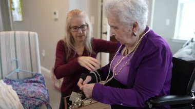 Tekerlekli sandalyedeki olgun bir kadına kıyafet seçen genç bir kadın yardımcı olur. İçerideler, kıyafet seçenekleriyle ilgili düşünceli bir sohbete giriştiler..