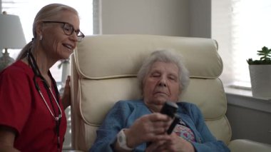 Önlüklü bir bakıcı, bir sandalyede oturan yaşlı bir kadınla iletişim kurar, ev gibi bir sağlık ortamında ilişki ve duygusal destek sağlar..