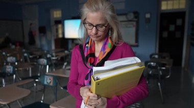 Olgun bir kadın boş bir sınıfta elinde dosyalarla derslerine hazırlanıyor. Onun terk edilmiş ifadesi onun üzüntüsünü ve öğretideki hayal kırıklığını yansıtıyor..