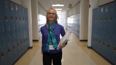 Profesyonel giyinmiş olgun bir kadın okul koridorunda elinde kitap ve kağıtlarla rahatça dolaşır. Durumunda samimiyet ve profesyonellik yayıyor..