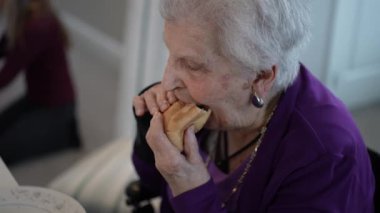 Yaşlı bir kadın masada otururken sandviçten büyük bir ısırık alır ve bir aile üyesi arka planda eşyaları düzenler ve sıcak bir atmosfer yaratır..
