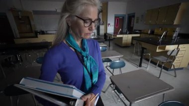 Berkeley Springs, WV, ABD - 11 25 2024: Profesyonel giyinmiş olgun kadın öğretmenin yakın çekimi, boş bir okul biyoloji laboratuvarında duruyor, elinde kitap tutuyor ve hazırlanırken üzgün ve mutsuz görünüyor