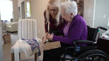 Bir bakıcı, tekerlekli sandalyedeki yaşlı bir kadına el işi projesinde yardım eder. Parlak ve misafirperver bir oturma odasında etkileşimlerini gösterir..