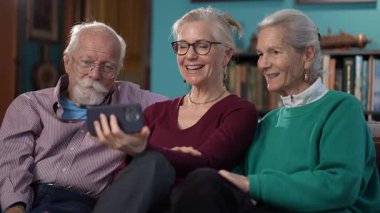 Yaşlı ebeveynler ve yetişkin kızlar, renkli oturma odalarında kitap ve dekor dolu gülücükler ve gülücükler sergileyen bir video görüşmesinde neşeli bir anı paylaşıyorlar..