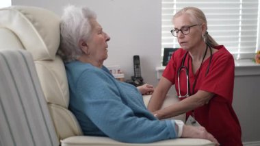 Kendini adamış bir bakıcı yaşlı bir kadınla ilgilenir, doğal ışıkla dolu rahat bir odada destek sunar, sağlık hizmetlerinde şefkatin önemini gösterir..