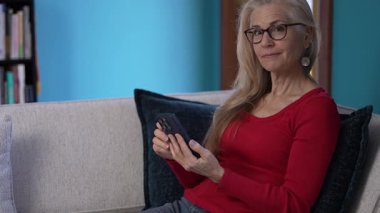 Uzun sarı saçlı ve gözlüklü olgun bir kadının portresi gülümsüyor, kameraya bakıyor, akıllı telefon tutuyor. Parlak mavi duvar sıcak atmosfere canlı bir dokunuş ekler..